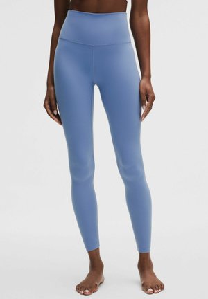 Femeie purtând colanți albastru deschis, mulați, cu talie înaltă, stând desculță pe un fundal alb simplu.