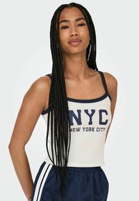 Top corto bianco con bordi blu navy, con la scritta "NYC New York City" stampata in caratteri in grassetto. Abbinato a pantaloni corti sportivi scuri e capelli lunghi intrecciati.