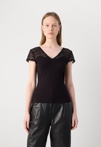 T-shirt noir à col en V avec des manches en dentelle, présentant un design ajusté et une texture lisse ; porté avec un pantalon en cuir noir taille haute.