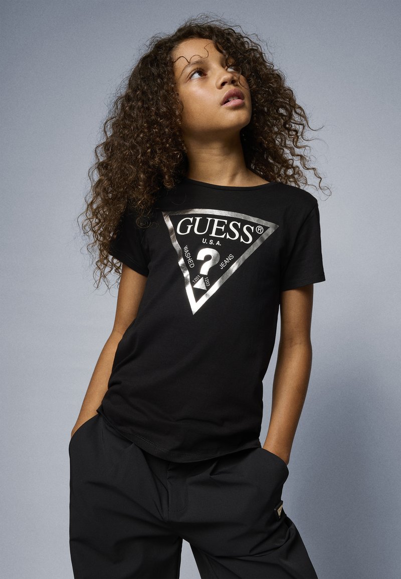 Black cotton t-shirt featuring a silver triangle design with "GUESS" and "?". Styled with relaxed black pants.