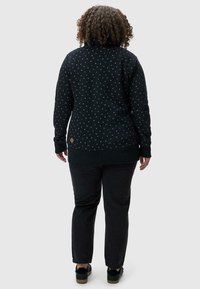 Suéter negro con un patrón texturizado de pequeños puntos de colores, que presenta un cuello alto y puños acanalados, combinado con pantalones oscuros y zapatos negros.
