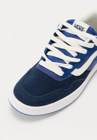 Baskets Vans bleu et marine avec lacets blancs, bande latérale blanche et semelle blanche sur un fond clair uni.