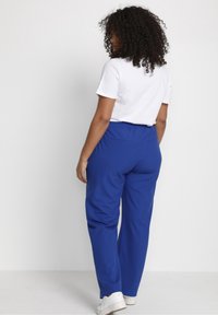 Blaue Hosen mit elastischem Bund und zwei Gesäßtaschen. Das Modell trägt ein weißes T-Shirt und weiße Turnschuhe, Ansichten von hinten.