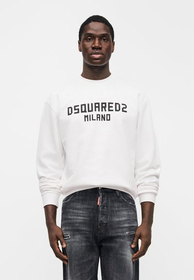 Άνδρας που φοράει λευκό φούτερ DSQUARED2 Milano και σκούρα ξεθωριασμένα τζιν, στέκεται μπροστά σε απλό φωτεινό φόντο.