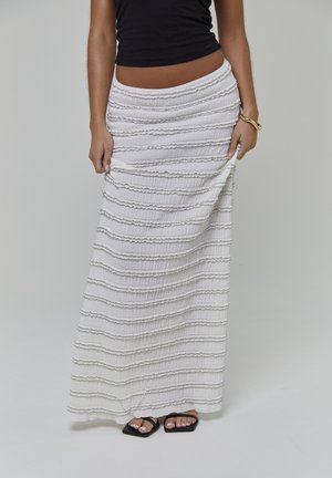 Witte maxi-rok met getextureerde, horizontale strepen en een geplooide tailleband. Gecombineerd met zwarte platte sandalen en een aansluitende zwarte top.