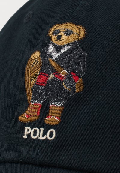 Melnā cepure ar izšūtu lāci formālajā tērpā ar saulesbrillēm, sarkaniem akcentiem un tekstu "POLO" zem tā.