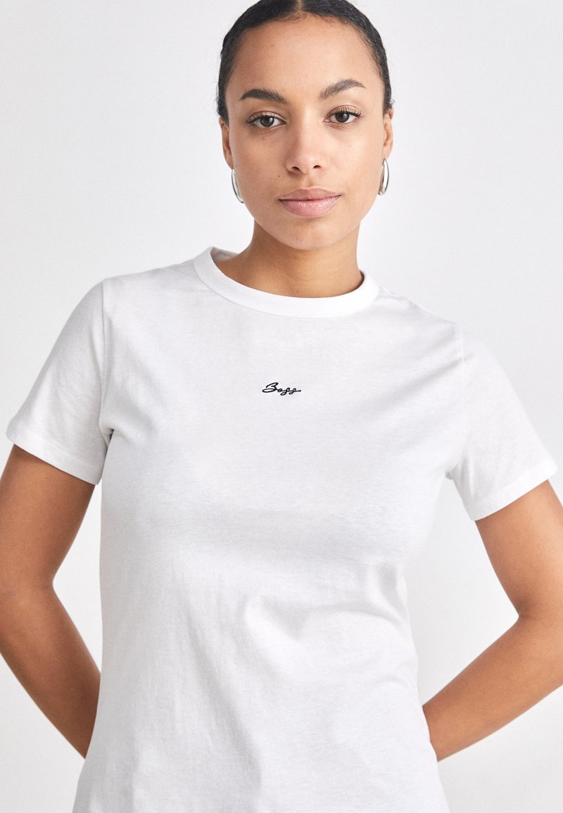 T-shirt en coton blanc à manches courtes, décolleté rond, avec un logo brodé noir au centre de la poitrine. Texture lisse, coupe décontractée.