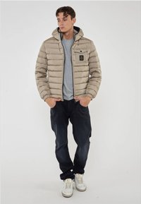 Giacca a vento beige con cappuccio, cuciture orizzontali e tasca sul petto. Indossata sopra una maglietta grigia e abbinata a jeans scuri e sneakers bianche.