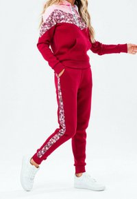 Mujer vestida con un hoodie burdeos con panel floral y joggers a juego, con zapatillas blancas, posando frente a un fondo blanco liso.