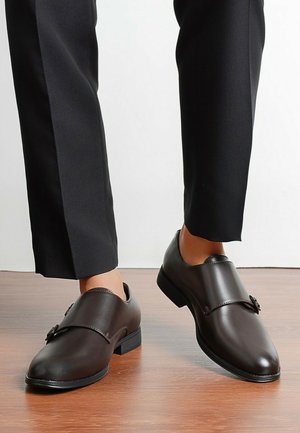 Chaussures habillées à sangle de moine en cuir marron portées avec un pantalon noir sur mesure sur un sol en bois.