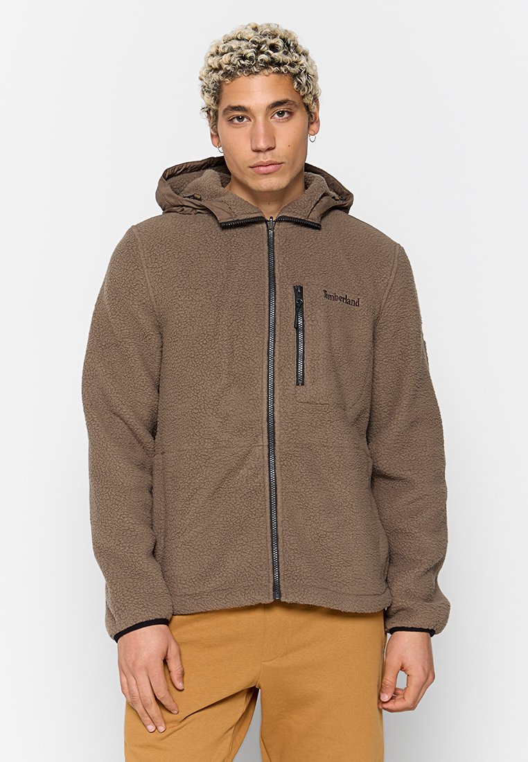 Brązowa polarowa bluza z kapturem na zamek błyskawiczny, z kieszenią zapinaną na zamek na klatce piersiowej oraz logo na górnej lewej stronie.