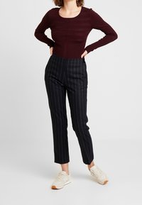 Haut ajusté bordeaux à manches longues avec des rayures texturées, associé à un pantalon sombre à fines rayures. Le mannequin porte des baskets claires.