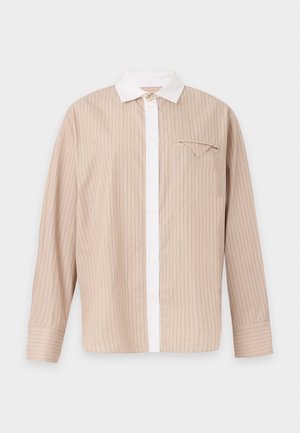 Femme aux cheveux bouclés, debout, portant une chemise beige à boutons, un pantalon large foncé et des talons ouverts noirs sur un fond clair uni.