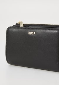 Crna kožna novčanica sa zatvaračem na patent, s brendom "BOSS" ispisanim zlatnim slovima. Glatka tekstura s urednim šavovima.