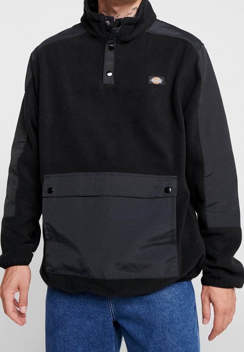 Chaqueta tipo pulóver negra de forro polar con cuello alto, cierre de botones a presión y un bolsillo frontal con detalle de botón.