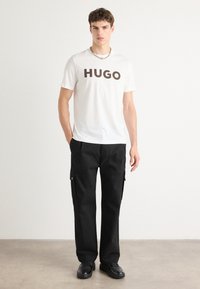 Fehér pamut póló sötétbarna "HUGO" logóval, fekete cargo nadrággal párosítva, zsebekkel és fém részletekkel.