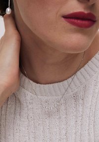 Srebrny naszyjnik z małymi okrągłymi perłami i zdobieniami, noszony nad jasnym, teksturowanym swetrem, blisko obszaru szyi.