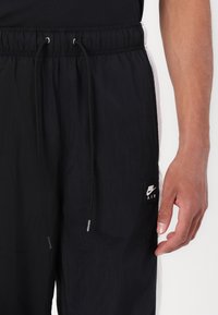 Man som bär svarta Nike Air träningsbyxor med vita nätpaneler på sidorna och svarta dragsnören, handen avslappnad vid sidan.