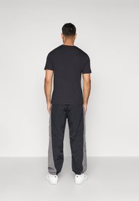 Man staat met zijn rug naar de camera, gekleed in een zwart t-shirt, zwart-grijze trainingsbroek en witte sneakers, tegen een effen grijze achtergrond.