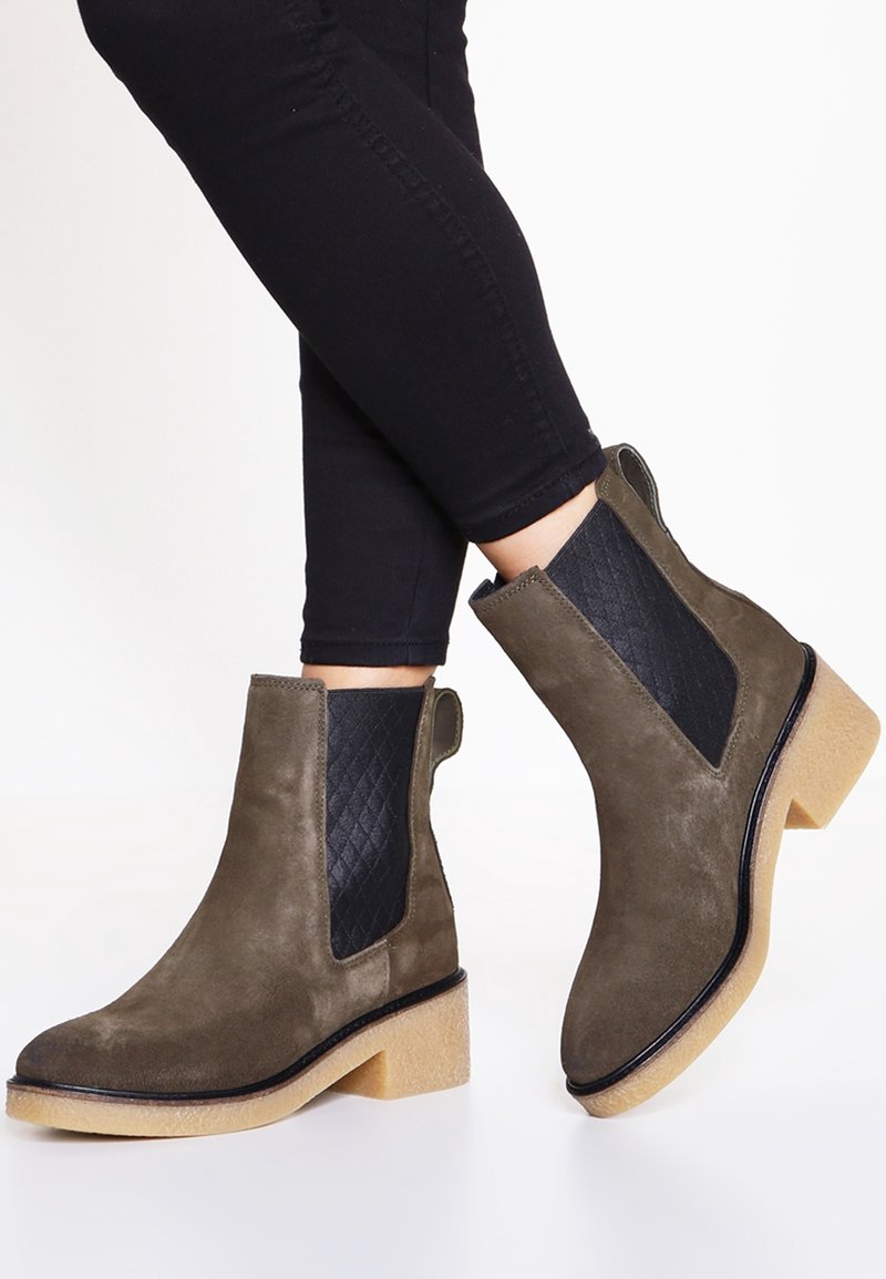 Femme portant des bottines Chelsea en daim vert olive avec des côtés élastiques noirs et des semelles en crêpe beige clair, associées à un pantalon noir ajusté.