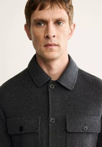 Abrigo de lana gris oscuro con un cuello clásico, que cuenta con dos bolsillos en el pecho, cierres de botones y un acabado de tejido texturizado.