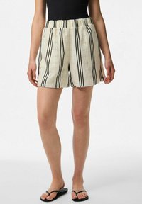 Pantaloni corti a strisce bianche con strisce verticali nere, vita elasticizzata, tessuto texturizzato e tasche laterali. Indossati con infradito nere.