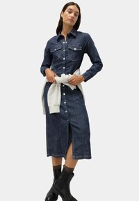 Denim klänning med långa ärmar, knappstängning framtill, två bröstfickor, mörkblå färg och en sidoklyvning, i kombination med en ljusgrå tröja knuten i midjan.