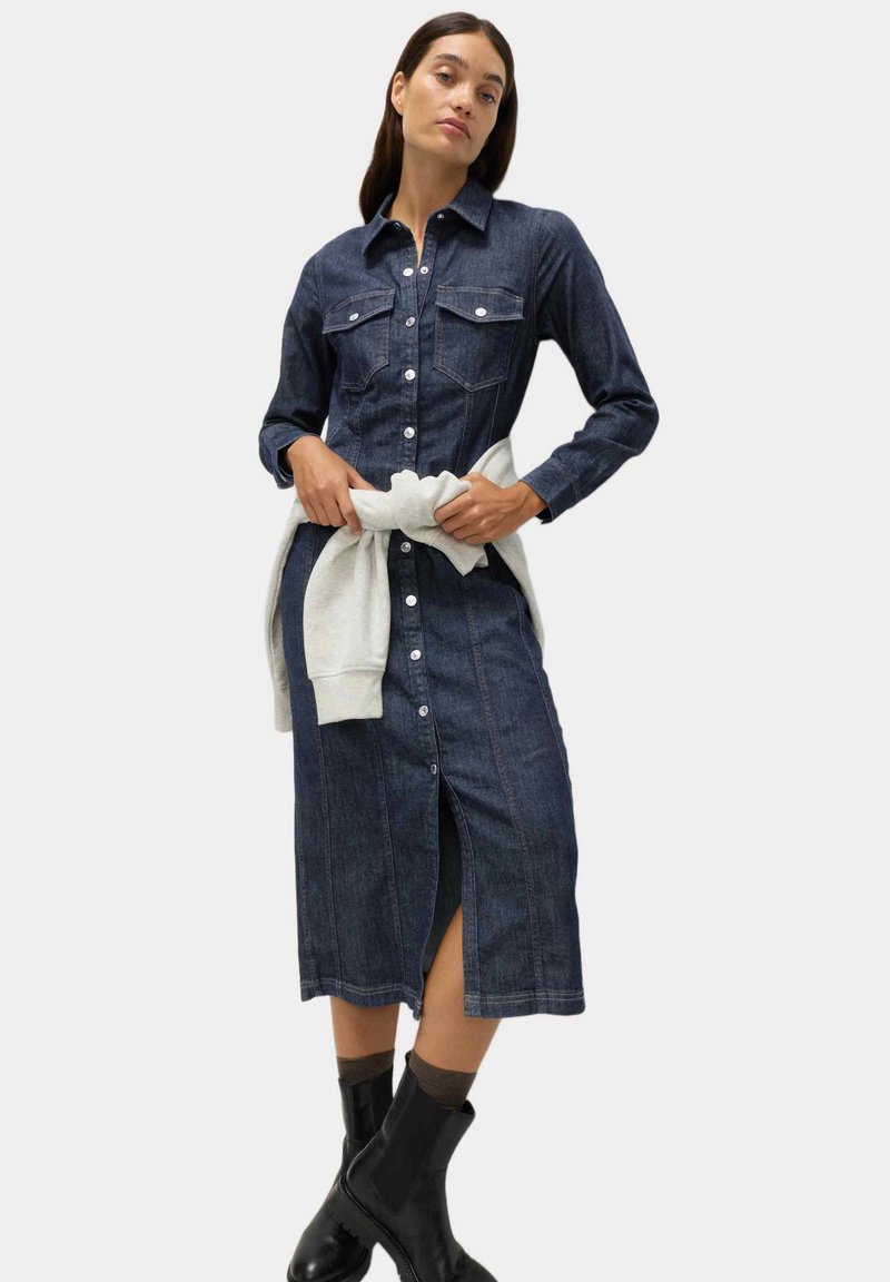 Vestido camisa de mezclilla con mangas largas, botones en el frente, dos bolsillos en el pecho, color azul oscuro y una abertura lateral, combinado con un suéter gris claro atado a la cintura.