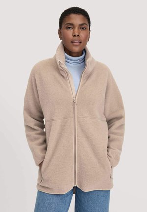 Fleece jack in licht beige, met een hoge kraag, voorkant rits en zijzakken. Draag het over een lichtblauwe coltrui, met een relaxte pasvorm.