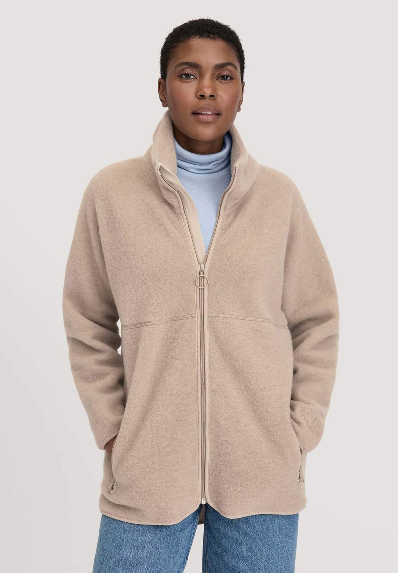 Giacca in pile di un beige chiaro, con colletto alto, zip frontale e tasche laterali. Indossata sopra un dolcevita azzurro chiaro, con una vestibilità comoda.