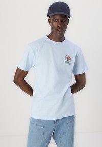 T-shirt de algodão azul claro com mangas curtas, apresentando um patch gráfico floral no lado esquerdo do peito. Combinado com jeans de denim azul claro.