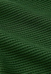 Tissu tricoté vert foncé avec un côtes ondulées texturées et de petites perforations rectangulaires créant un motif répétitif.