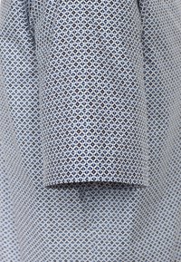 Das Shirt-Gewebe weist ein geometrisches Muster in Marineblau und Hellblau auf, mit kleinen Rautenformen und einer glatten Textur. Nahaufnahme des Ärmel-Details.