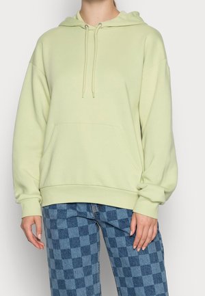 Kobieta w jasnzie zielonym oversize'owym kapturze z przednią kieszenią i niebieskich, w kratę dżinsach, stojąca na jednolitym tle.