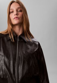 Jonge vrouw met steil lichtbruin haar draagt een zwarte leren jas met rits tegen een effen grijze achtergrond.