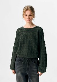Vrouw in een donkergroene gestructureerde gebreide trui met lange mouwen en zwarte high-waisted jeans, staand tegen een witte achtergrond.