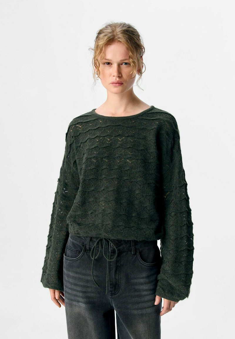 Vrouw in een donkergroene gestructureerde gebreide trui met lange mouwen en zwarte high-waisted jeans, staand tegen een witte achtergrond.