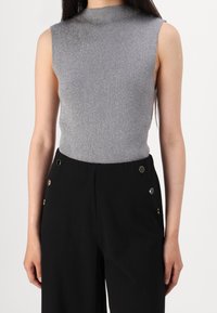 Zilveren geribbelde mouwloze top met een hoge hals, gecombineerd met zwarte wijde broek met gouden knopen aan de zijkant.