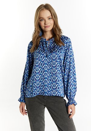 Langärmlige blaue Bluse mit Paisleymuster, mit gerafftem Ausschnitt und Rüschenärmeln, aus leichtem Stoff. Getragen mit grauen Hosen.