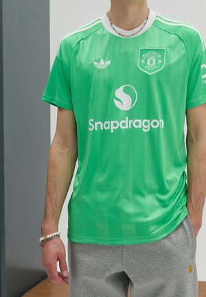 Person, die ein grünes Manchester United Fußballtrikot mit Snapdragon-Logo und grauen Jogginghosen trägt, steht drinnen vor grauen und weißen Wänden.
