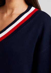 Marineblauer Strickpullover mit V-Ausschnitt, aus strukturiertem Stoff und einem gestreiften Kragen in Rot, Weiß und Marineblau.