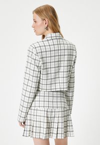 Kuratblazer und plissierter Rock aus weißem Stoff mit schwarzem kariertem Muster. Verfügt über lange Ärmel und ein schlichtes Kragendesign.