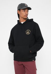 Sweatshirt preto com capuz, bolso frontal e um design de logotipo dourado. Usado com calças bordeaux, acompanhado por um boné cinza.