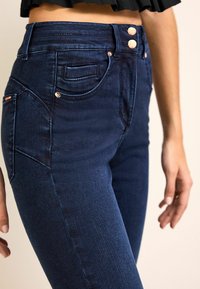 Jeans de tiro alto en azul marino con una textura suave, que presentan dos botones en la parte delantera, costuras sutiles y un pequeño bolsillo para monedas en el lado izquierdo.