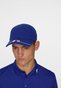 Blå basebollkeps i tyg med en böjd skärm och ett vitt zick-zack-mönster längs kanten. Tillskuren design med matchande blå skjorta.