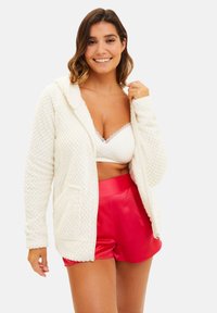 Haut à capuche texturé crème avec une capuche, des poches avant et une fermeture éclair intégrale. Porté par-dessus un bralette blanc et associé à un short rouge brillant.
