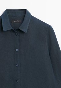 Granatowa, lniana koszula z długim rękawem, kołnierzykiem i pięcioma guzikami z przodu, pokazująca górną część tułowia oraz metkę marki wewnątrz kołnierzyka.