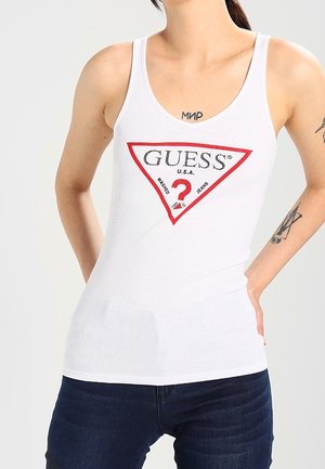 Person iført en hvid tanktop med et rødt og sort Guess-logo og mørkeblå jeans, der viser tatoveringer på arme og øvre bryst.