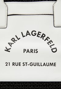 Väsk i vitt läder med svart text som säger "KARL LAGERFELD PARIS 21 RUE ST-GUILLAUME." Har sömmar och strukturerade handtag.