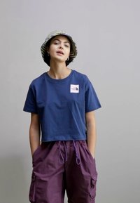 Navy cropped t-shirt with a white logo on the left chest, paired with purple cargo shorts. Model wears a checkered bucket hat.